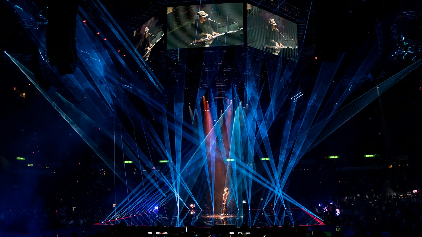 Arena stage with sweeping blue laser beams and elevated IMAG screens above a performer