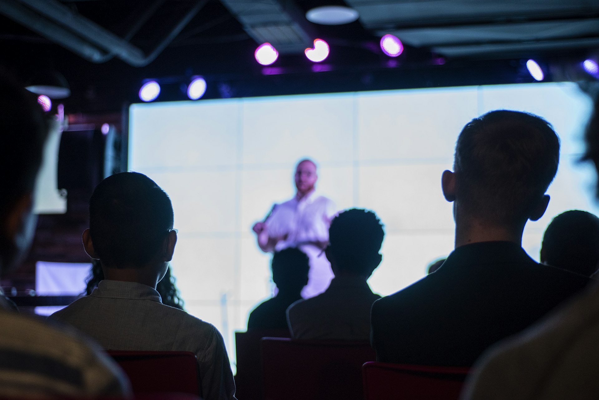 Seated audience watching a speaker on a large tiled LED video wall in a dark room