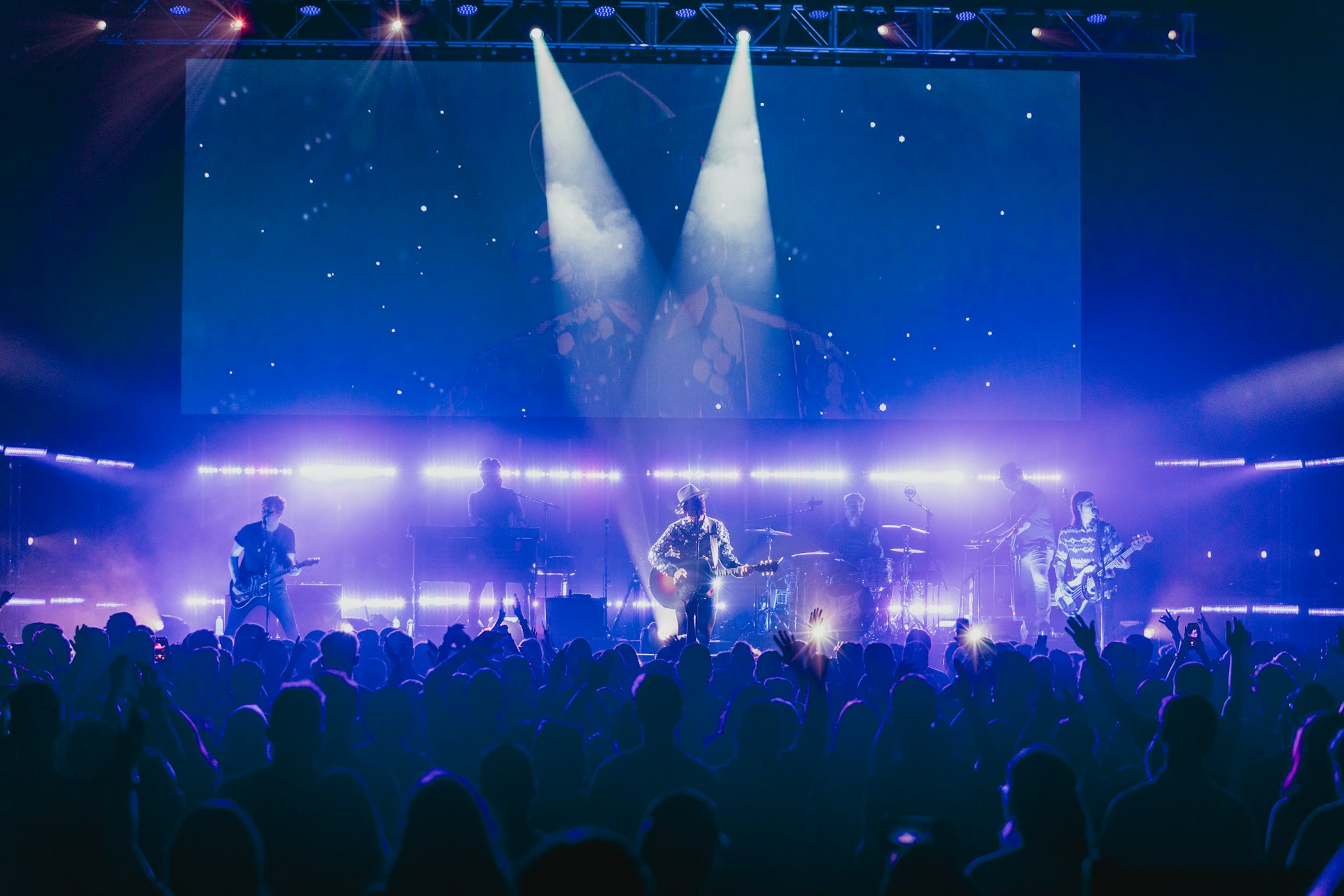 Band performing on a concert stage in front of a large LED screen backdrop with blue beams and a packed crowd