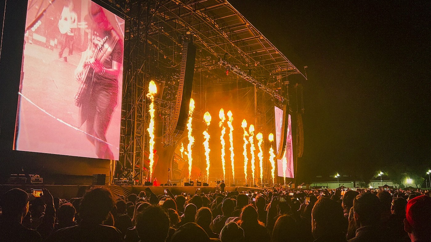Outdoor night concert with large LED side screens, flame jets and a crowd in front of the stage